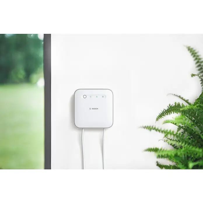 Bosch Smart Home Controller II - ZigBee 3.0, Weiß/Silber - Nahaufnahme eines Bosch Smart Home Controllers mit Kabeln an einer weißen Wand, neben einem Fenster und einer Pflanze.