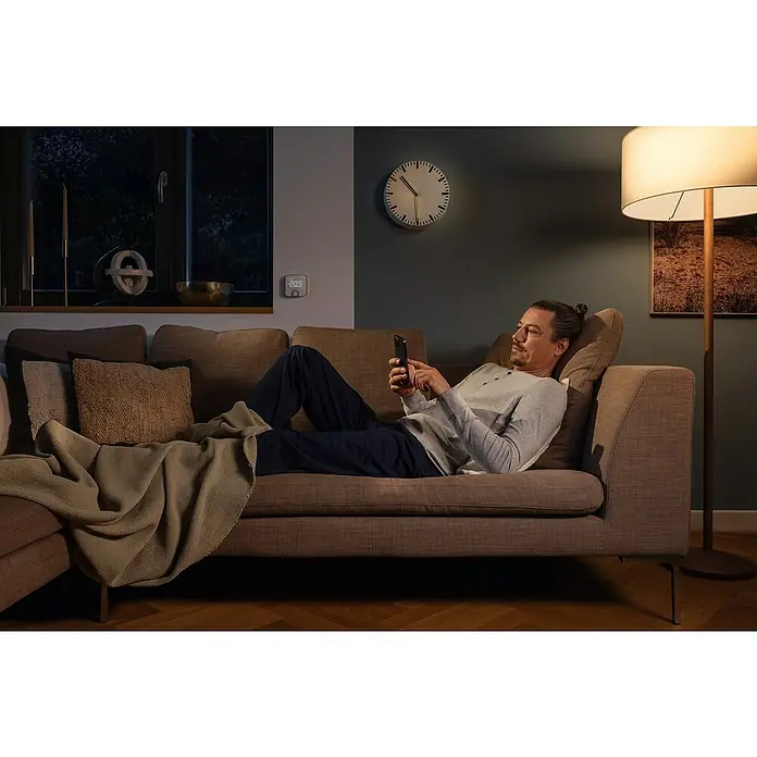 Bosch Smart Home Raumthermostat II - Weiß, Silber, L x B x H: 87 x 86 x 35 mm - Ein Mann entspannt auf der Couch und bedient sein Smartphone. Im Hintergrund sind eine Uhr und eine Stehlampe zu sehen.