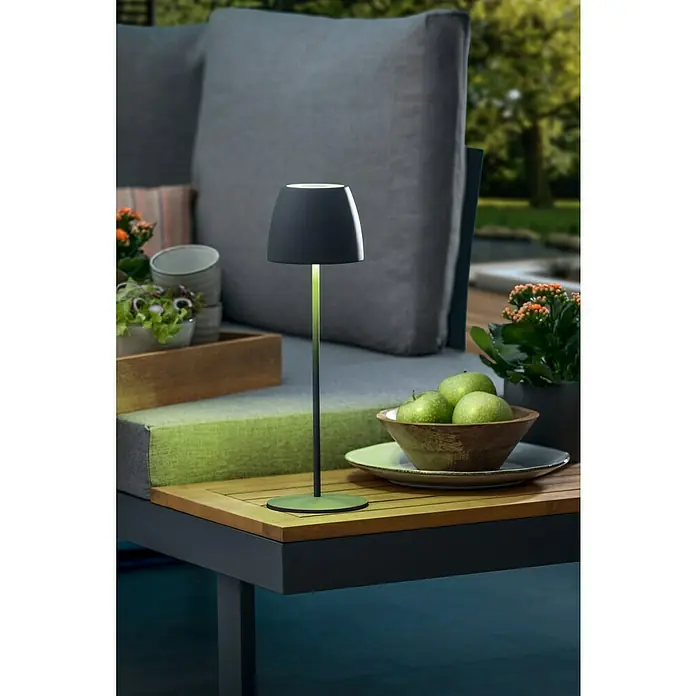 Lavida Lámpara de sobremesa a batería Silea - 3,3 W, Antracita, Color de luz: RGBW - Un primer plano muestra una moderna lámpara de mesa colocada sobre una mesa de madera junto a un cuenco de manzanas verdes, ofreciendo un ambiente cálido.