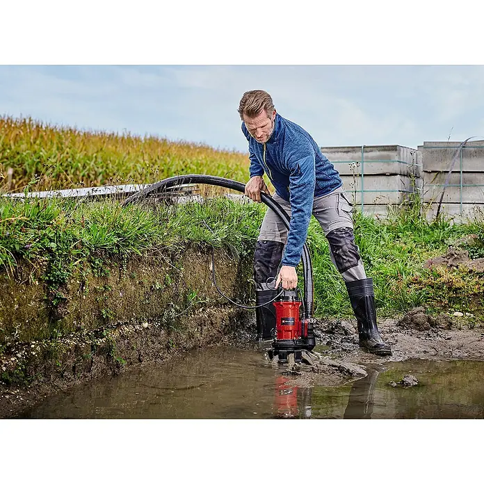 Einhell Bomba para aguas residuales GE-DP 900 Cut - 900 W, Capacidad de bombeo máx.: 16.000 l/h, Profundidad de inmersión: 7 m - Un hombre opera una bomba sumergible para aguas sucias Einhell roja en una zanja fangosa, rodeada de vegetación y losas de hormigón.