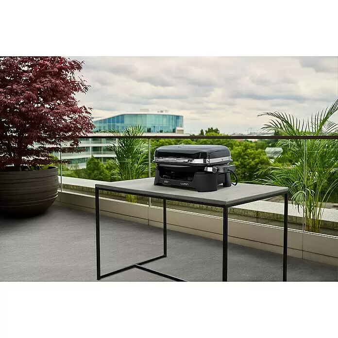 Weber Barbacoa eléctrica Lumin 2000 - Superficie de la parrilla (An x Pr): 49 x 33 cm, 2,2 kW, Negro - Parrilla eléctrica Weber Lumin negra sobre una mesa gris de exterior en un balcón, rodeada de plantas y vistas a la ciudad.