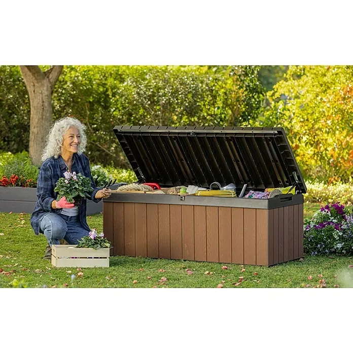 Baúl para jardín Darwin 100 - L x An x Al: 65 x 143 x 55 cm, Marrón - Una mujer se arrodilla junto a una caja de almacenamiento para exteriores marrón con la tapa abierta, llena de herramientas de jardinería. Ideal para guardar.