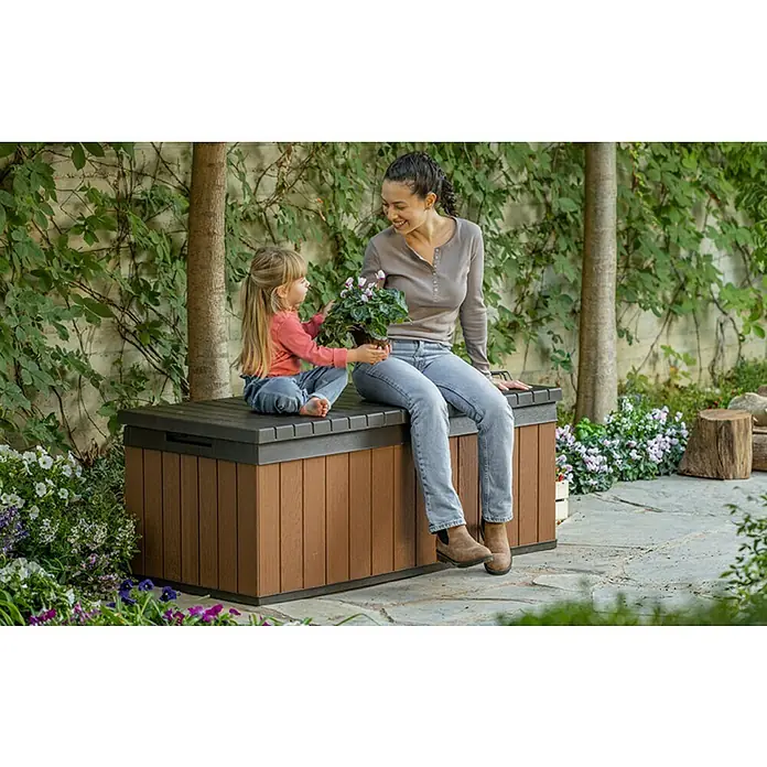 Baúl para jardín Darwin 100 - L x An x Al: 65 x 143 x 55 cm, Marrón - Banco marrón de exterior con una mujer y una niña sentadas en él, rodeado de exuberante vegetación. El banco proporciona asientos cómodos al aire libre.