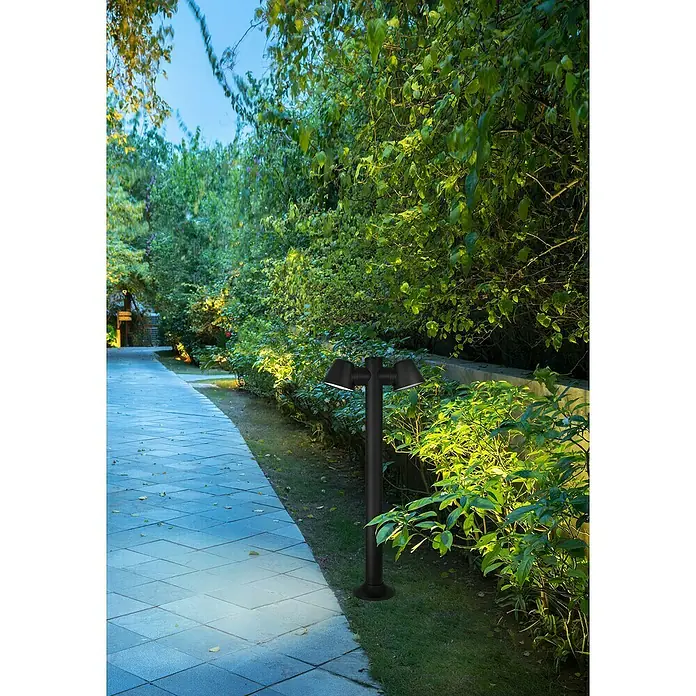 Forlight Cone Lámpara de pie para exterior - 16 W, Color del marco: Negro, 11,7 x 15 x 80 cm, Sin bombillas - Una moderna lámpara de jardín negra ilumina un camino pavimentado, rodeado de exuberante vegetación, emitiendo una luz cálida.