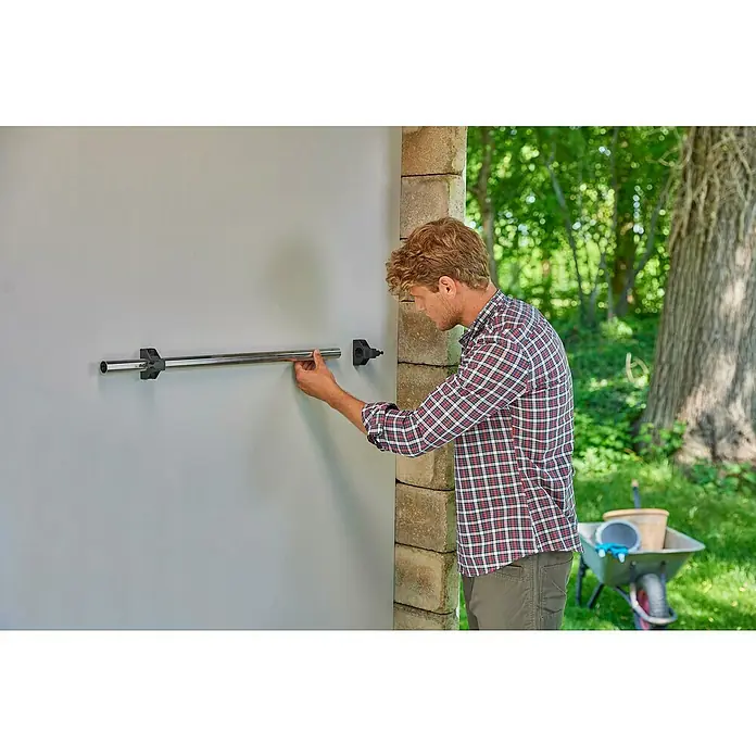 Gardena Halterung Plus - Traglast: 10 kg - Mann montiert eine verchromte Stange mit Halterungen an einer grauen Wand, Werkzeugwagen mit Zubehör im Hintergrund.