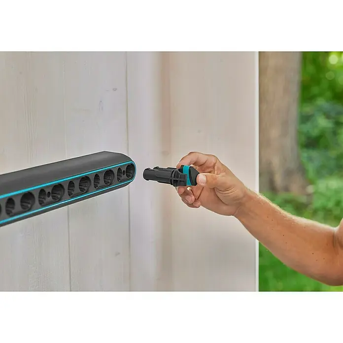 Gardena Halterung Flex - 10 kg, Kunststoff - Eine Person schließt ein schwarzes Gartengeräte-Zubehör an eine Halterung mit blauen Akzenten an einer weißen Wand an.