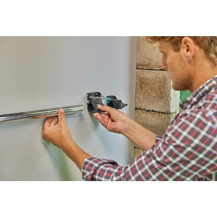 Gardena Halterung Set Einzelhalter & Einzelhaken - Traglast: 10 kg, Aluminium - Ein Mann befestigt eine silberne Stange mit einer schwarzen Halterung an einer grauen Wand. Schraubendreher wird verwendet.