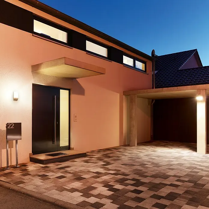 Steinel Sensor-Außenleuchte L 1 S schwarz - 60 W, 120 x 110 x 255 mm, Schwarz, IP44 - Außenaufnahme eines modernen Hauses mit stilvoller Beleuchtung, einem Carport und einer gepflasterten Auffahrt in der Dämmerung.