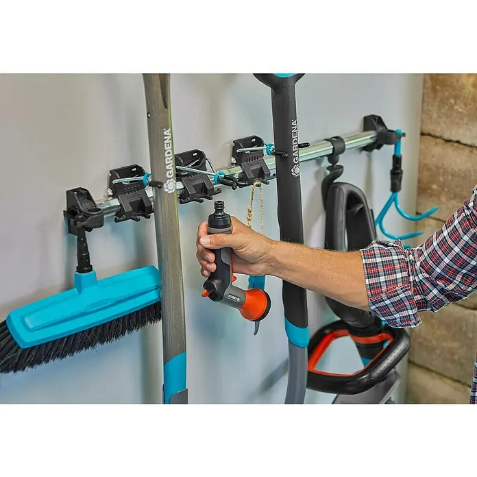 Gardena Halterung Plus - Traglast: 10 kg - Gardena Wandhalter mit diversen Gartengeräten wie Besen, Schaufeln und einer Spritzpistole, die von einer Hand gehalten wird.