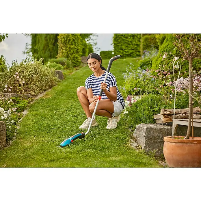 Gardena ComfortCut Tijeras cortacésped de batería Li Set - 3,6 V, 1 batería, Anchura de la hoja: 8 cm - Una mujer se agacha en el jardín, usando unas tijeras de césped inalámbricas Gardena con mango para recortar los bordes del césped, rodeada de flores.