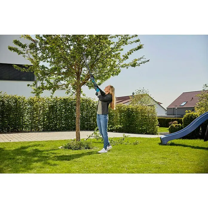 Gardena Power for All 18V Cortarramas de jardín - 18 V, Sin batería, Grosor de rama máx.: 32 mm - Una mujer poda un árbol en su jardín con las tijeras de podar GARDENA, rodeada de césped y setos, bajo un cielo azul.