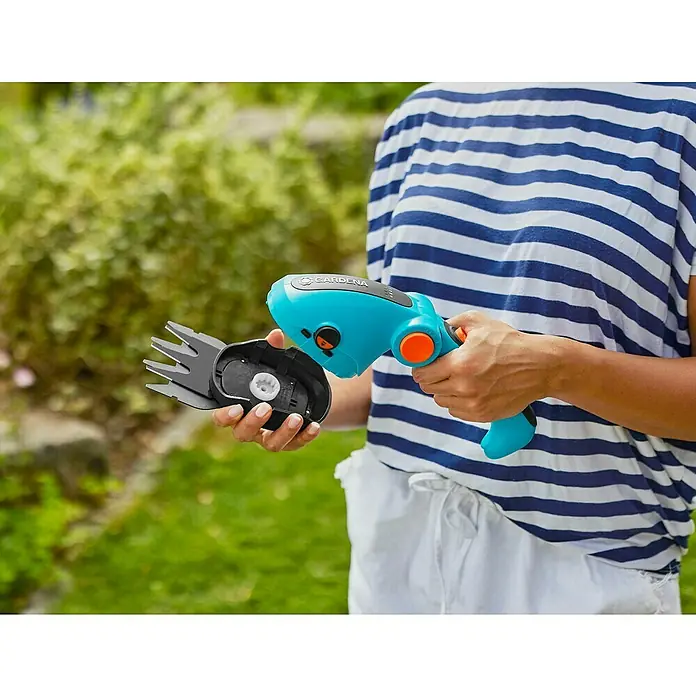 Gardena ComfortCut Podadora de batería Li - 3,6 V, Iones de litio, 1 batería, Anchura de la hoja: 8 cm - Una persona sostiene unas tijeras de césped inalámbricas Gardena en el jardín. El dispositivo tiene mango azul y accesorio de corte negro.