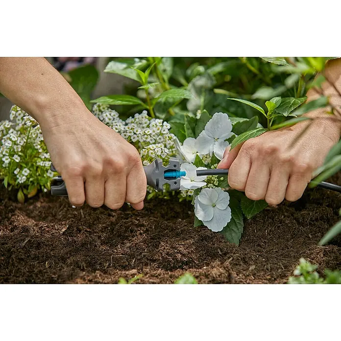 Gardena Micro-Drip Vorstecher - Passend für: Gardena Micro-Drip Artikel & Lochung des Verlegerohres, Kunststoff, Schwarz - Nahaufnahme einer Person, die ein Bewässerungssystem mit einem grauen Verbinder und einem blauen Einsatz in einem Garten mit Blumen installiert.