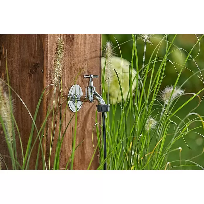 Gardena Micro-Drip Hahnanschlussstück - 3/16" x 4,6 mm - Verchromter Außenwasserhahn an einem Holzpfosten mit Schlauchanschluss, umgeben von grünen Gartenpflanzen.