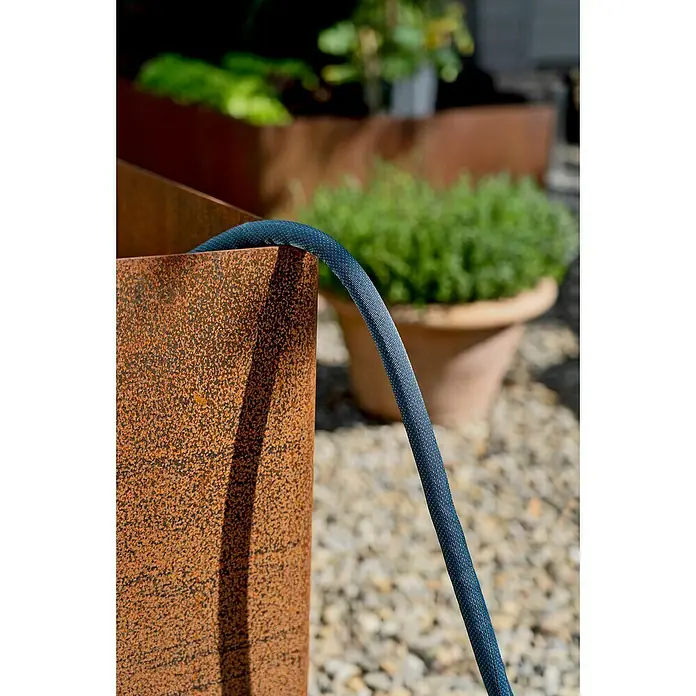 Gardena Gartenschlauch Liano Xtreme Set - Schlauch, Wasserstop, Schlauchverbinder, 2x Hahnverbinder, Bewässerungsbrause - Rostfarbener Pflanzkasten mit einem blauen Schlauch, der aus dem Kasten ragt, und einem grünen Busch im Hintergrund.