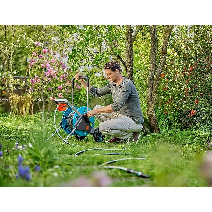 Gardena EcoLine Gartenschlauch Set - 20 m, Ausstattung: Anschlussstücke - Ein Mann hockt im Garten und benutzt eine blaue Gardena Schlauchbox mit Rädern, um seinen Rasen zu bewässern.