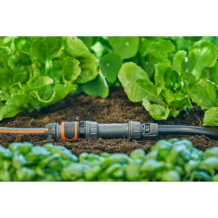 Gardena Micro-Drip Hahnanschlussstück - 1/2" x 13 mm - Detailaufnahme einer Gardena-Schlauchverbindung, die zwischen zwei Schläuchen im Gartenbeet liegt, umgeben von frischem Grün.