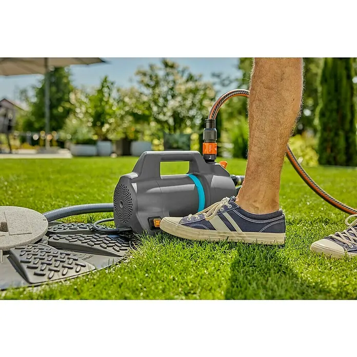 Gardena Gartenpumpen-Set 4100 Silent Saugschlauch550 W, Max. Fördermenge: 4.100 l/h - Eine Gardena Gartenpumpe liegt auf dem Rasen neben einer Revisionsöffnung, während ein Mann in blauen Sneakern im Garten steht.