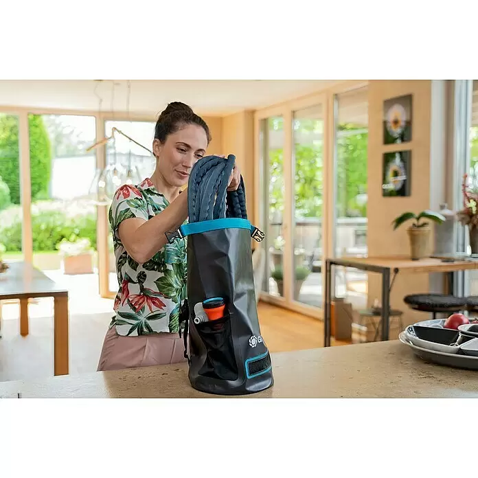 Gardena Gartenschlauch Liano Xtreme Set mit Hahnverbinder & Aufbewahrungstasche - 7 -tlg., Kunststoff - Eine Frau steckt einen aufgerollten blauen Schlauch in eine dunkelgraue Tragetasche mit blauem Rand und Netztaschen, die Bewässerungsanschlüsse enthält.
