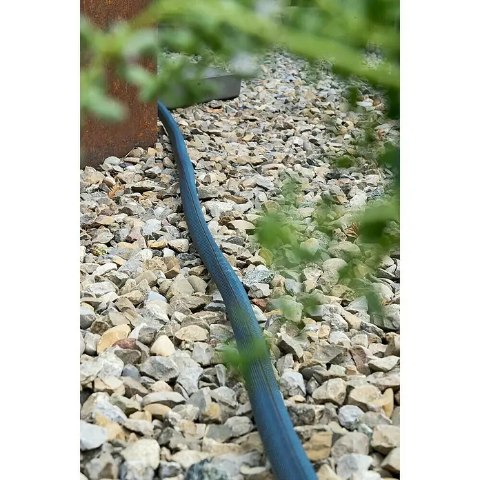 Gardena Gartenschlauch Liano Xtreme Set - 25 m, Schlauchdurchmesser: 19 mm - Ein blauer Bewässerungsschlauch liegt auf einem Kiesweg, teilweise verdeckt von grünen Pflanzen, ideal für Gartenbewässerung.