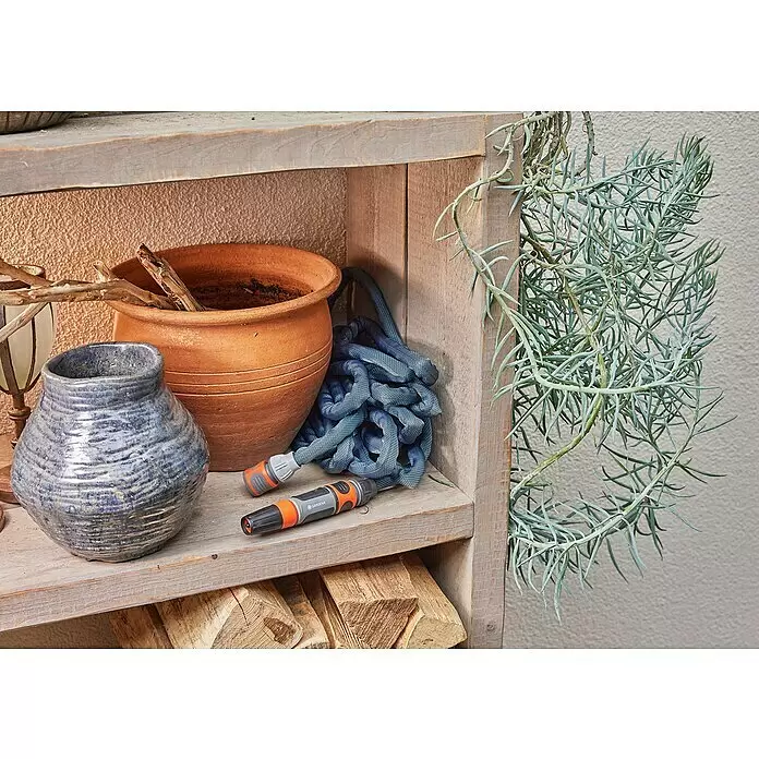 Gardena Gartenschlauch Liano Xtreme Set - 20 m, Schlauchdurchmesser: 13 mm - Ein blauer Textilgartenschlauch mit orangefarbenen und grauen Gardena-Anschlüssen, auf einem Holzregal neben einem Terrakottatopf und Brennholz.