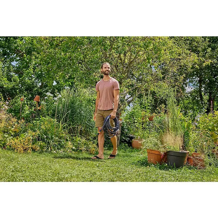 Gardena Gartenschlauch Liano Life Set - 15 m, Schlauchdurchmesser: 13 mm - Ein Mann, der mit einem Gartenschlauch in der Hand durch einen üppigen Garten geht. Der Schlauch ist dunkel und wirkt robust.