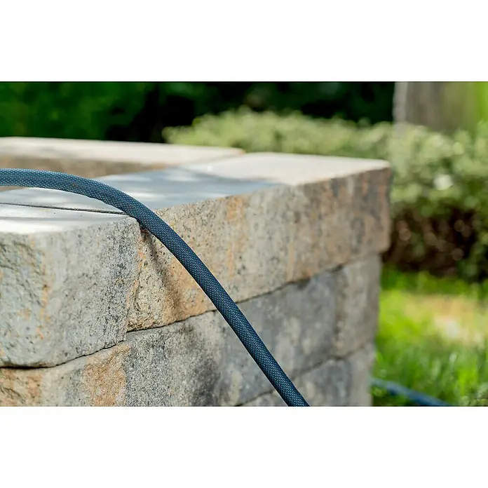 Gardena Gartenschlauch Liano Xtreme Set - Schlauch, Wasserstop, Schlauchverbinder, 2x Hahnverbinder, Bewässerungsbrause - Nahaufnahme eines blauen Gartenschlauchs, der auf einer gestapelten Steinmauer liegt, mit üppigem Grün im Hintergrund.