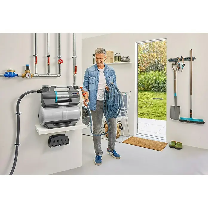 Gardena Hauswasserwerk 5600 Silent Comfort - 650 W, Max. Fördermenge: 5.600 l/h, Max. Druck: 4,7 bar - Ein Mann hält einen Schlauch neben einer Gardena Hauswasseranlage mit Zubehör in einem Hauswirtschaftsraum.