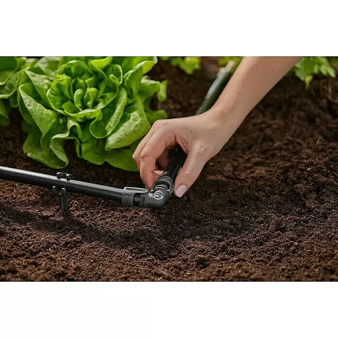 Gardena Micro-Drip L-Stück - 1/2" x 13 mm, 2 Stk. - Hand verbindet schwarzes Bewässerungsrohr mit grauem Winkelstück in Gartenerde neben grünem Salat.