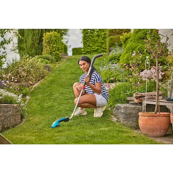 Gardena ClassicCut Podadora de batería - 3,6 V, Iones de litio, 1 batería, Anchura de la hoja: 8 cm - Mujer en cuclillas en el jardín utilizando unas tijeras de césped inalámbricas GARDENA para bordes de césped precisos. Perfecto para jardinería sin esfuerzo.