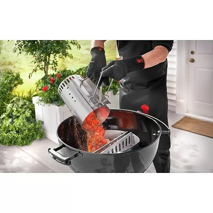 Weber Anzündkamin Rapidfire - Ø x H: 19 x 30 cm - Eine Person in schwarzen Handschuhen gießt glühende Holzkohle aus einem silbernen Anzündkamin in einen schwarzen Grill, dabei sprühen Funken.