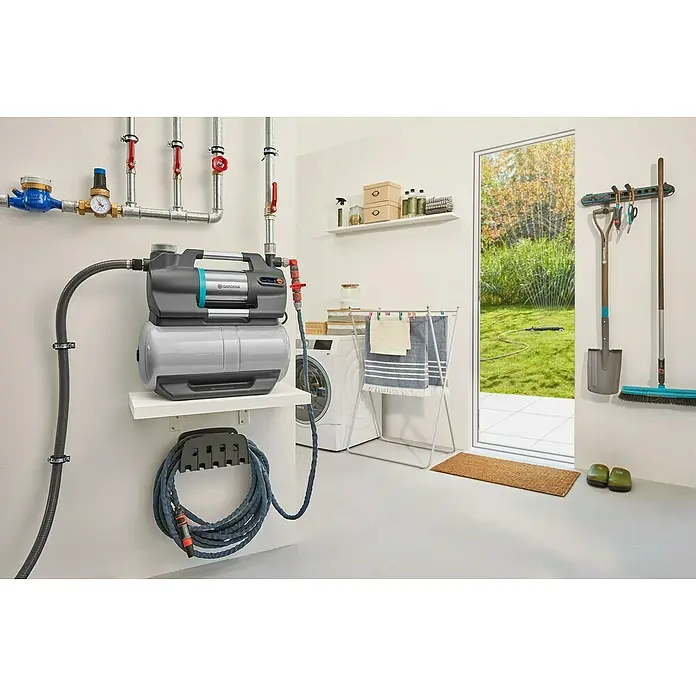 Gardena Hauswasserwerk 5600 Silent Comfort - 650 W, Max. Fördermenge: 5.600 l/h, Max. Druck: 4,7 bar - Das Bild zeigt eine Gardena Hauswasseranlage mit Zubehör, montiert in einem Hauswirtschaftsraum, mit einem Schlauch und einer Pumpe auf einer Ablage.