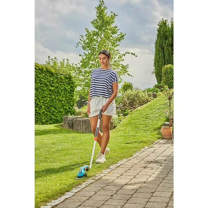 Gardena ComfortCut Tijeras cortacésped de batería Li Set - 3,6 V, 1 batería, Anchura de la hoja: 8 cm - Una mujer utiliza unas tijeras de césped a batería con mango telescópico para recortar el césped a lo largo de un camino pavimentado en un jardín.
