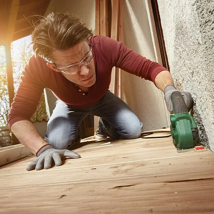 Bosch NanoBlade-Säge AdvancedCut 50 - 500 W - Ein Mann kniet und benutzt eine grüne Bosch Schleifmaschine, um eine Holzbodenfläche zu bearbeiten. Er trägt Schutzbrille und Handschuhe.