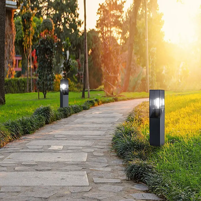 Forlight Munic Lámpara de pie para exterior - 15 W, Color del marco: Antracita, 13 x 13 x 40 cm, Sin bombillas - Elegantes luces de jardín bordean un camino de piedra, iluminando el paso a través del paisaje verde mientras el sol de la tarde crea un ambiente cálido.
