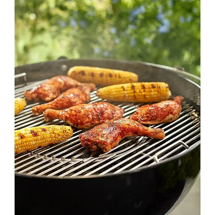 Weber Gourmet BBQ System Grillrost - L x B: 56,61 x 54,61 cm, Durchmesser: 57 cm, Verchromter Stahl - Gegrillte Hähnchenkeulen und Maiskolben auf einem Holzkohlegrill mit grüner Natur im Hintergrund.