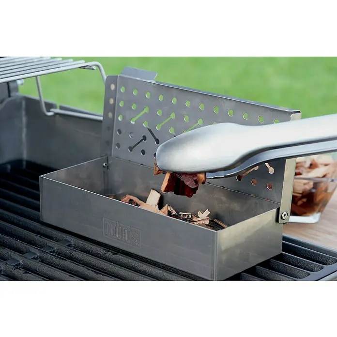 Weber Chips ahumadores - Específico para: Ahumar aves, 700 g - La imagen muestra una caja ahumadora Weber para parrillas de carbón, en la que se están colocando astillas de ahumado con pinzas.