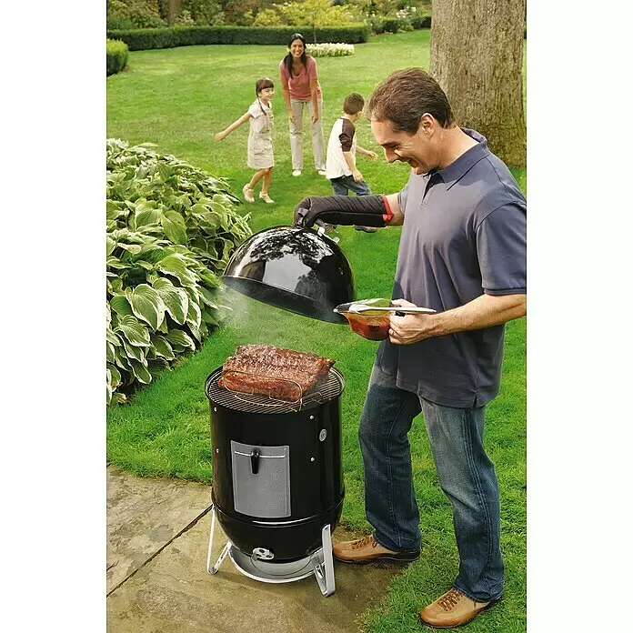 Weber Ahumador Mountain Cooker - Diámetro de la superficie de la parrilla: 47 cm, Negro - Un hombre cocina costillas en una parrilla ahumadora cilíndrica negra al aire libre, con un cepillo para untar y salsa.