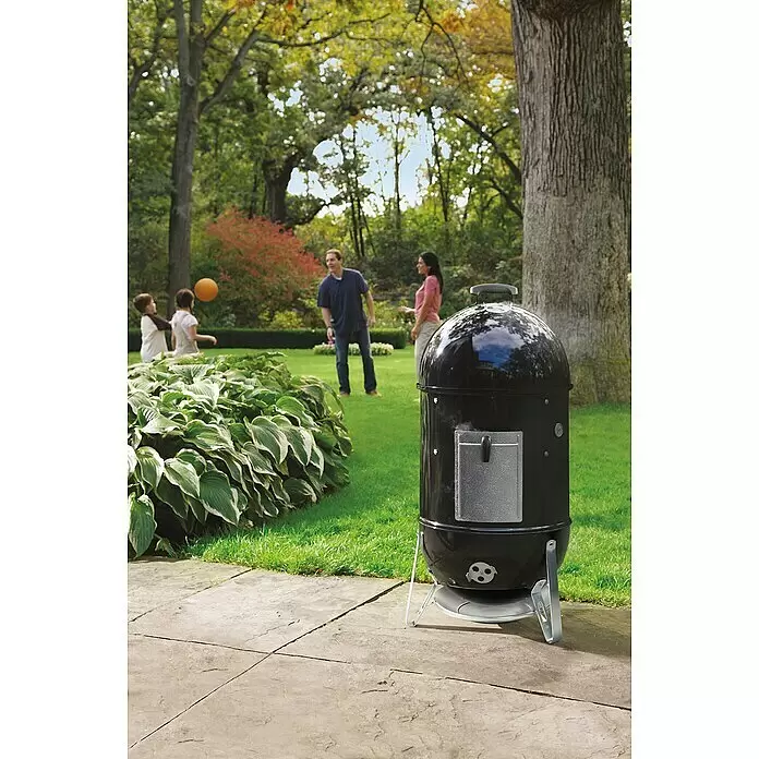 Weber Ahumador Mountain Cooker - Diámetro de la superficie de la parrilla: 47 cm, Negro - Ahumador de carbón negro con puerta de acceso y asa plateadas, en un patio trasero con una familia al fondo.