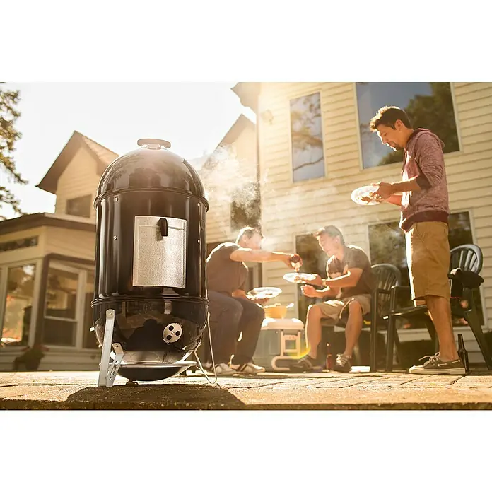 Weber Räucherofen Smokey Mountain CookerDurchmesser Grillfläche: 47 cm, Schwarz - Schwarzer, aufrechter BBQ-Smoker im Freien mit drei Männern im Hintergrund. Ein entspanntes Barbecue mit Freunden.