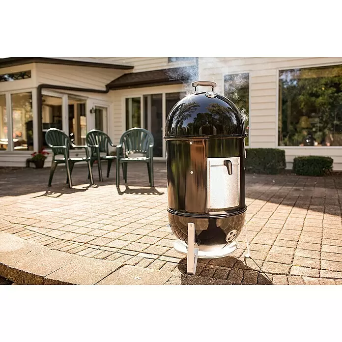 Weber Ahumador Mountain Cooker - Diámetro de la superficie de la parrilla: 47 cm, Negro - Ahumador negro con detalles plateados, echando humo activamente en un patio junto a una casa con sillas.