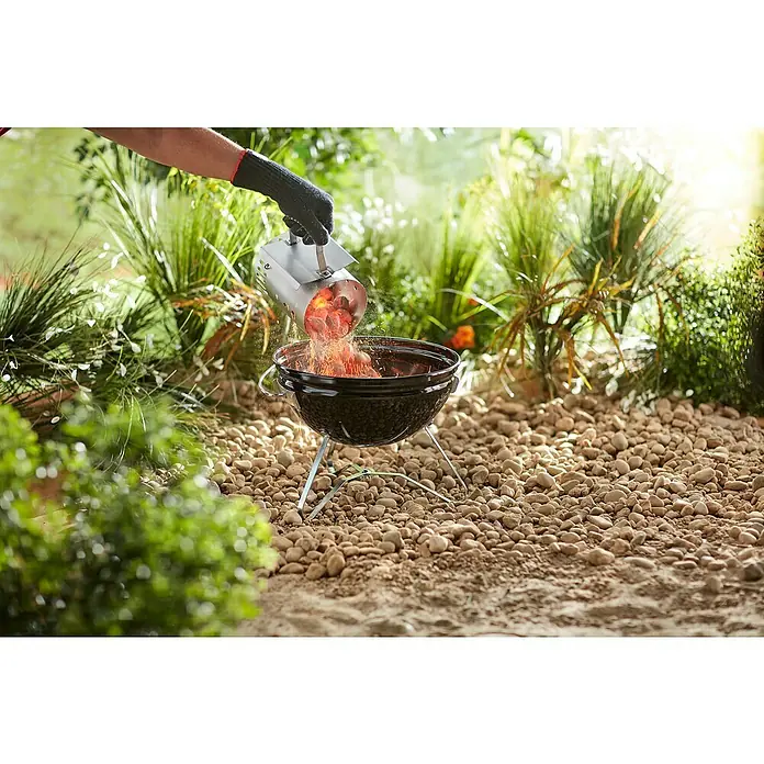 Weber Anzündkamin - B x T x H: 14,5 x 26,5 x 20,5 cm - Eine Person leert mit einem Anzündkamin glühende Holzkohle in einen schwarzen Kugelgrill. Im Hintergrund ist eine Gartenlandschaft zu sehen.