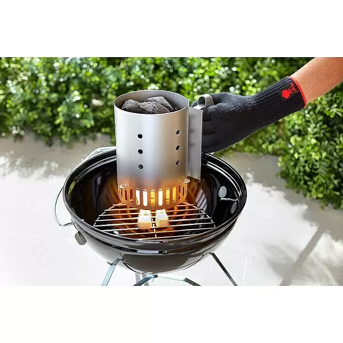 Weber Anzündkamin - B x T x H: 14,5 x 26,5 x 20,5 cm - Eine Hand im schwarzen Handschuh hält einen silbernen Anzündkamin mit schwarzer Holzkohle über einem schwarzen Grill, Flammen sind unten sichtbar.