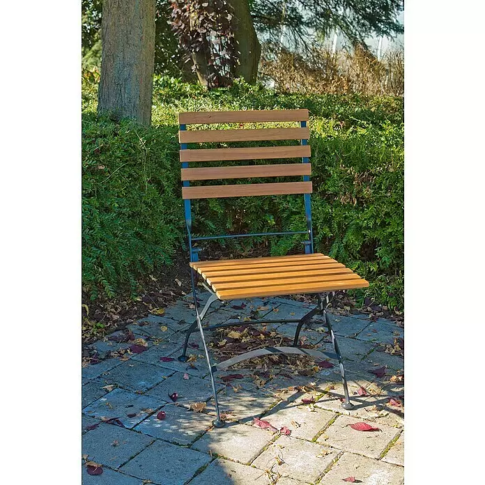 Acamp Gartenmöbel-Set Lindau - 3 -tlg., Akazie, Eisen, Braun, Klappbar, mit und ohne Armlehne - Ein Klappstuhl mit hellbraunen Holzlatten und dunklem Metallgestell steht auf einer gepflasterten Terrasse, umgeben von grünem Laub.