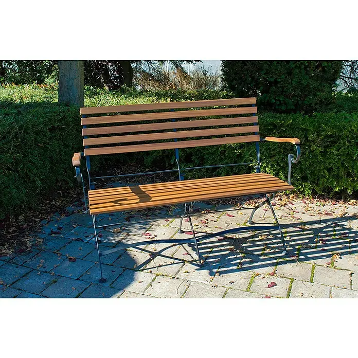 Acamp Gartenmöbel-Set Lindau - 4 -tlg., Akazie, Eisen, Braun, Stuhl ohne Armlehne - Holzbank im Freien auf einem gepflasterten Bereich mit Schatten, umgeben von Grün. Die Bank lädt zum Verweilen im Garten oder Park ein.