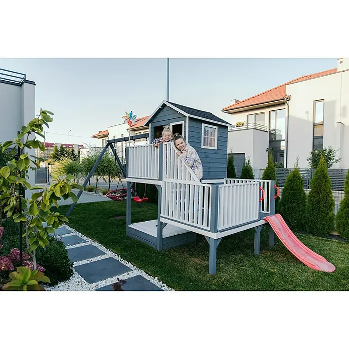 Kinderspielturm Tom - Natur - Kinder spielen auf einem blaugrauen Holzspielhaus mit weißem Geländer, einer Rutsche und Schaukeln in einem üppigen Garten.