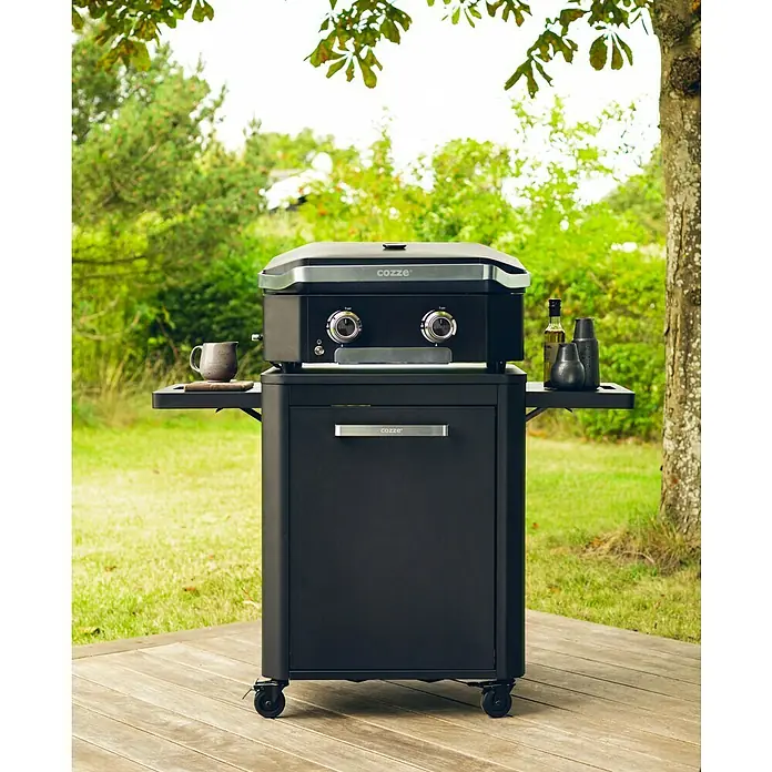 Cozze Unterschrank - L x B x H: 60 x 124 x 83 cm, Schwarz - Schwarzer Cozze-Grill mit Unterschrank auf einer Holzterrasse, umgeben von Grün. Perfekt für Grillabende im Freien.