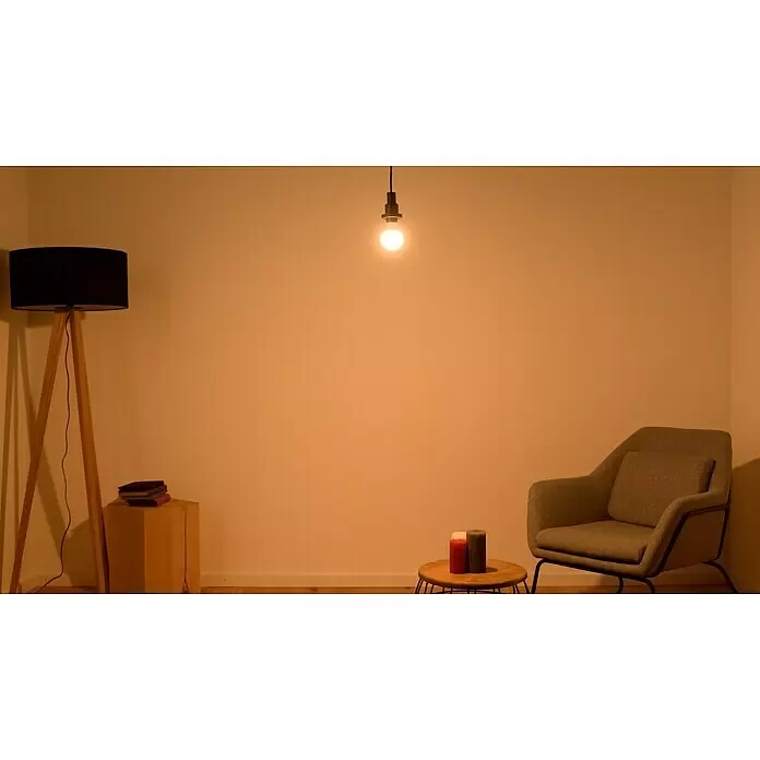 Osram LED-Lampe Glühlampenform E27 matt - E27, Nicht Dimmbar, Warmweiß, 806 lm, 8,5 W - Gemütliches Wohnzimmer mit Holz-Dreibein-Stehlampe, hängender Glühbirne, grauem Sessel, Holzschemel mit Büchern und kleinem Tisch mit Kerzen.