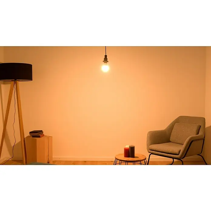 Osram LED-Lampen Set Glühlampenform E27 matt - E27, Nicht Dimmbar, Warmweiß, 1.521 lm, 13 W, 3 Stk. - Gemütliches Wohnzimmer mit Stehlampe, Sessel und Kerzen auf kleinem Tisch, beleuchtet durch eine hängende Glühbirne. Warme und einladende Atmosphäre.
