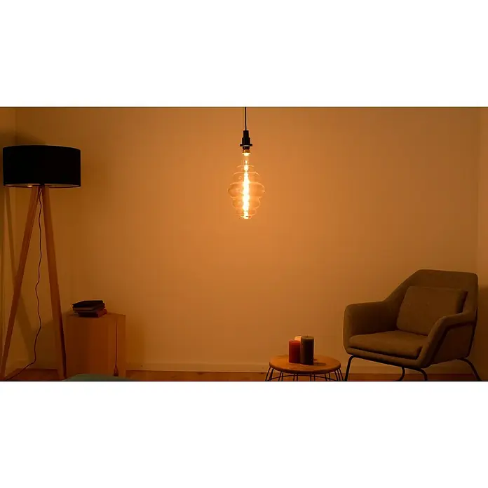 Osram LED-Lampe Vintage Edition 1906 Glühlampenform E27 - E27, Dimmbar, Warmweiß, 360 lm, 4,8 W - Die Glühbirne hängt von der Decke und beleuchtet das Zimmer mit warmem Licht, neben einer Stehlampe, einem Sessel und Kerzen auf einem kleinen Tisch.
